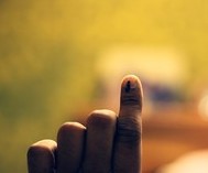image of an index finger with indelible ink on the nail, which is proof that a person has voted in India. The finger is held up, and the background is yellow and blurred